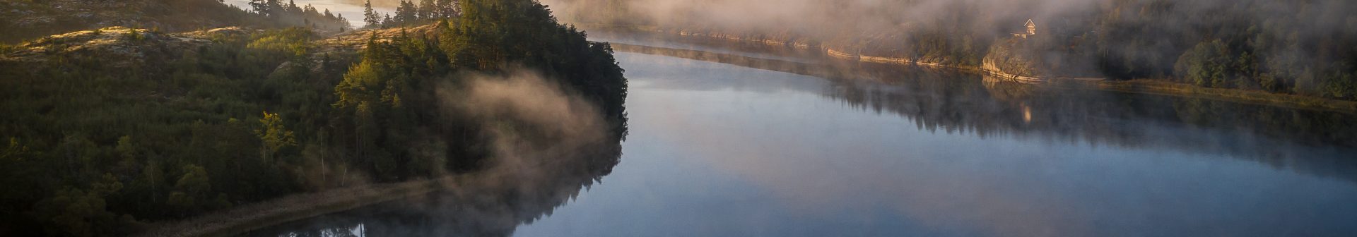 Dimma svävar över en sjö omgiven av skogsklädda kullar i gryningen.