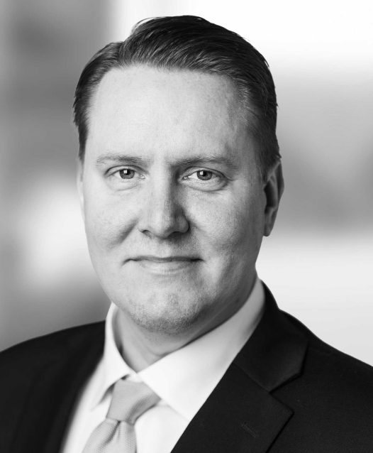 A black and white portrait of a man in a suit and tie, smiling slightly, with a blurred background.